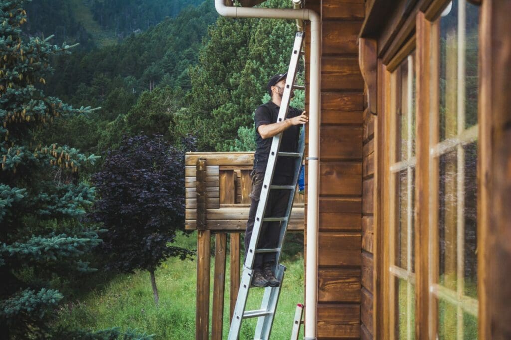 Man on a ladder working on a wooden cottage with trees and mountains